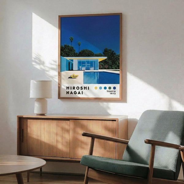 May include: Framed poster displaying modern architectural illustration of minimalist house with swimming pool, palm trees, and bright blue sky, hanging above wooden sideboard with table lamp, next to mid-century modern green upholstered armchair in contemporary living room setting with natural lighting casting shadows on white wall.