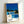 May include: Framed art print leaning against white wall showing mid-century modern house with swimming pool under bright blue sky. Modernist architecture features glass walls, flat roof with yellow accent, surrounded by palm trees and lush greenery. Yellow lounge chairs sit on white pool deck beside crystal blue water. Clean geometric design with vibrant blues, yellows, and greens in retro illustration style. Light wood frame displays artwork at an angle on floor.