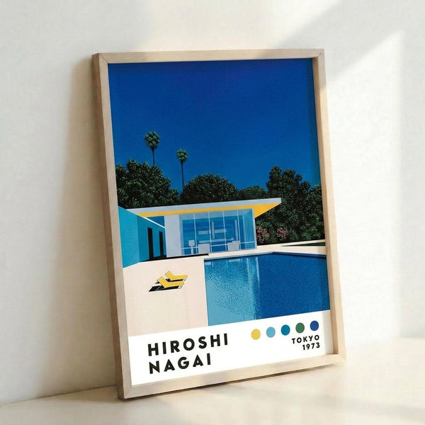 May include: Framed art print leaning against white wall showing mid-century modern house with swimming pool under bright blue sky. Modernist architecture features glass walls, flat roof with yellow accent, surrounded by palm trees and lush greenery. Yellow lounge chairs sit on white pool deck beside crystal blue water. Clean geometric design with vibrant blues, yellows, and greens in retro illustration style. Light wood frame displays artwork at an angle on floor.