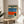 May include: Framed poster print displayed on round wooden side table next to beige upholstered chair in sunlit room with plant. Artwork shows collage-style composition with poolside scene featuring blue water, orange geometric shape, person wearing white headphones among rocks, and lounge chairs on deck. Natural wood frame, soft shadows from window light.