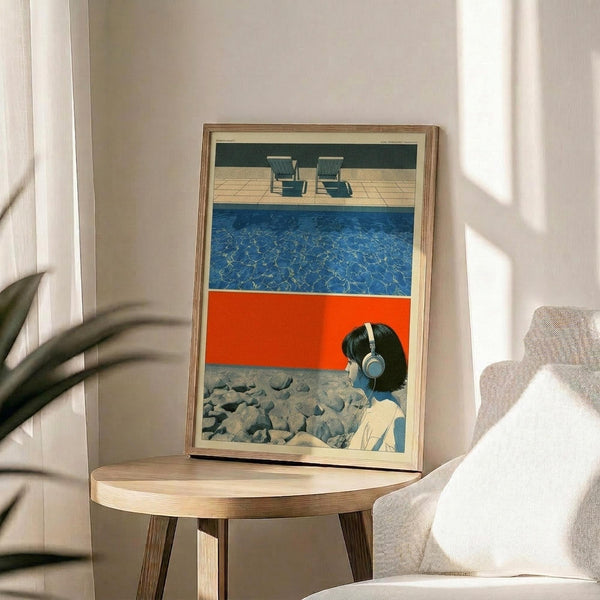 May include: Framed poster print displayed on round wooden side table next to beige upholstered chair in sunlit room with plant. Artwork shows collage-style composition with poolside scene featuring blue water, orange geometric shape, person wearing white headphones among rocks, and lounge chairs on deck. Natural wood frame, soft shadows from window light.