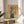 May include: Framed vintage-style poster with yellow background featuring woman in blue tones holding Coca-Cola bottle, red Japanese text overlay, displayed on round wooden side table next to beige upholstered chair with white pillow, plant leaves visible in corner, natural lighting creating soft shadows on white wall