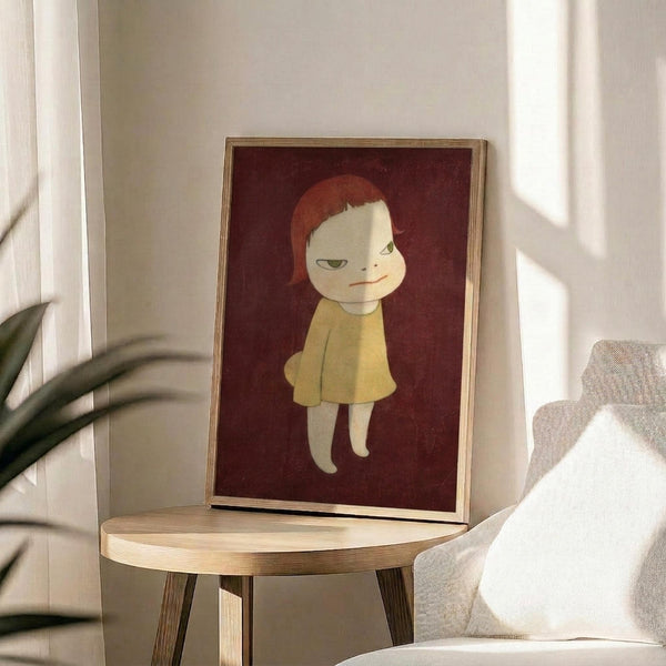 May include: Framed art print featuring a stylized illustration of a pale child figure with reddish-orange hair wearing a yellow dress against a deep burgundy background, displayed on a light wood round side table next to a beige upholstered chair with white pillow, green plant leaves visible, natural sunlight streaming across white walls creating soft shadows in minimalist modern living room setting.