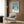 May include: Light wood framed poster showing swimming pool scene with palm trees and blue sky, displayed on wood paneled wall above cream upholstered armchair with geometric patterned pillow. Modern brass floor lamp and dark round side table with open book complete the neutral-toned living room setting.