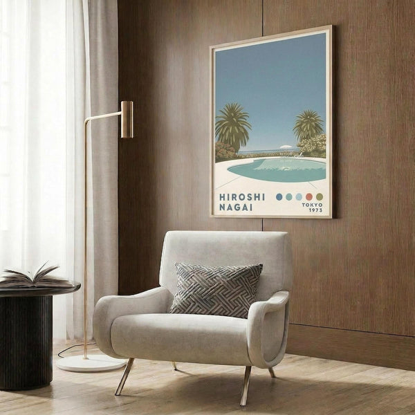 May include: Light wood framed poster showing swimming pool scene with palm trees and blue sky, displayed on wood paneled wall above cream upholstered armchair with geometric patterned pillow. Modern brass floor lamp and dark round side table with open book complete the neutral-toned living room setting.