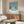 May include: Mid-century modern living room featuring a framed poster of a swimming pool scene with palm trees and blue sky. The artwork hangs above a wooden credenza with brass hardware, alongside a white curved chair, brass floor lamp, and glass-top coffee table. The room has beige walls and natural lighting creating warm shadows across the wooden furniture.