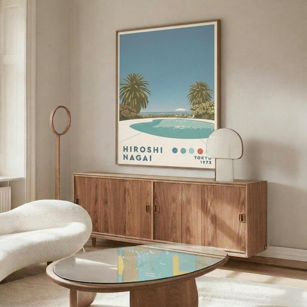 May include: Mid-century modern living room featuring a framed poster of a swimming pool scene with palm trees and blue sky. The artwork hangs above a wooden credenza with brass hardware, alongside a white curved chair, brass floor lamp, and glass-top coffee table. The room has beige walls and natural lighting creating warm shadows across the wooden furniture.