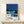 May include: Framed poster showing modern white house with swimming pool under bright blue sky, surrounded by palm trees and lush greenery. Mid-century architectural illustration in vibrant blues, yellows and greens with "HIROSHI NAGAI" and "TOKYO 1973" text below. Natural wood frame displayed on light wooden surface with small wrapped gift beside it against cream wall.