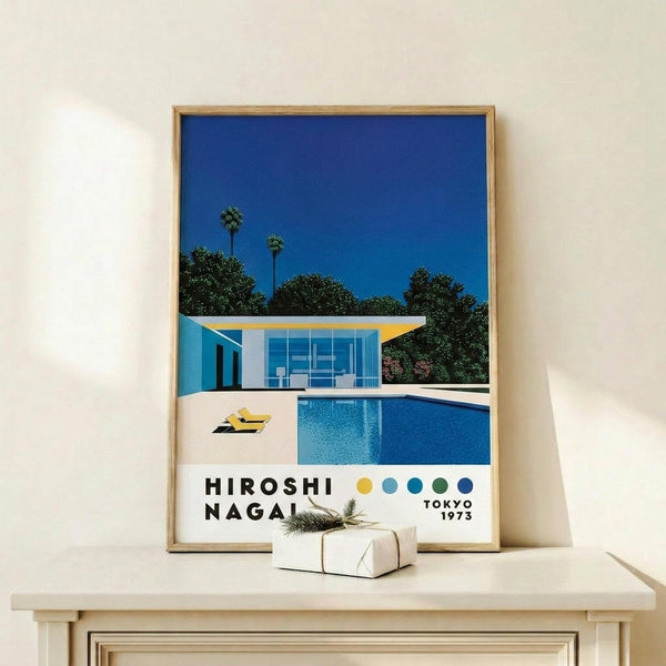 May include: Framed poster showing modern white house with swimming pool under bright blue sky, surrounded by palm trees and lush greenery. Mid-century architectural illustration in vibrant blues, yellows and greens with "HIROSHI NAGAI" and "TOKYO 1973" text below. Natural wood frame displayed on light wooden surface with small wrapped gift beside it against cream wall.