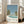 May include: Framed poster print leaning against beige sofa in bright white room with large windows. Art depicts tropical pool scene with two palm trees, turquoise swimming pool, mountain view, and blue sky in vintage travel poster style. Text shows "HIROSHI NAGAI" and "TOKYO 1973" with colorful dots. Natural wood frame, positioned on light wooden floor.