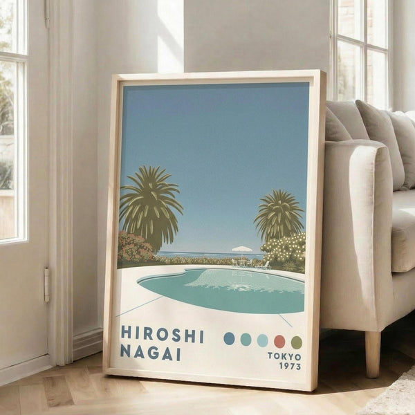 May include: Framed poster print leaning against beige sofa in bright white room with large windows. Art depicts tropical pool scene with two palm trees, turquoise swimming pool, mountain view, and blue sky in vintage travel poster style. Text shows "HIROSHI NAGAI" and "TOKYO 1973" with colorful dots. Natural wood frame, positioned on light wooden floor.