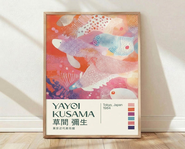 May include: Framed art poster leaning against white wall on light wooden floor. The artwork features abstract colorful whales or fish shapes in coral, pink, teal and purple with dotted patterns and flowing organic forms. Text reads "YAYOI KUSAMA Tokyo, Japan 1984" with Japanese characters below. Natural wood frame with clean modern aesthetic. Soft shadows cast on textured white wall behind.