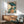 May include: Japanese ukiyo-e style artwork showing a woman in colorful kimono holding a teal umbrella, surrounded by large blue-green leaves. The print is displayed in a thin wooden frame on a white wall above a mid-century modern wooden credenza with sliding doors. A small white ceramic table lamp sits on the credenza, and a gray upholstered armchair with wooden frame is positioned nearby. Warm natural sunlight creates geometric shadows across the scene.