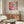 May include: Colorful abstract art poster with flowing organic shapes in pink, orange, blue and white featuring dotted patterns, displayed above a mid-century modern walnut credenza in a minimalist living room with cream walls, white upholstered chair, brass floor lamp, and glass-top coffee table