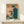May include: Framed vintage-style poster featuring Japanese text and illustration of person at retro computer workstation in orange, teal and navy blue color scheme, displayed on light wood surface alongside small wrapped gift box with twine and dried botanical accent against white wall