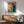 May include: Framed vintage-style poster with Japanese text and illustration of person using retro computer, displayed above wooden sideboard with white table lamp. Mid-century modern living room setting with sage green upholstered chair, round coffee table, and natural sunlight creating shadows on white wall.