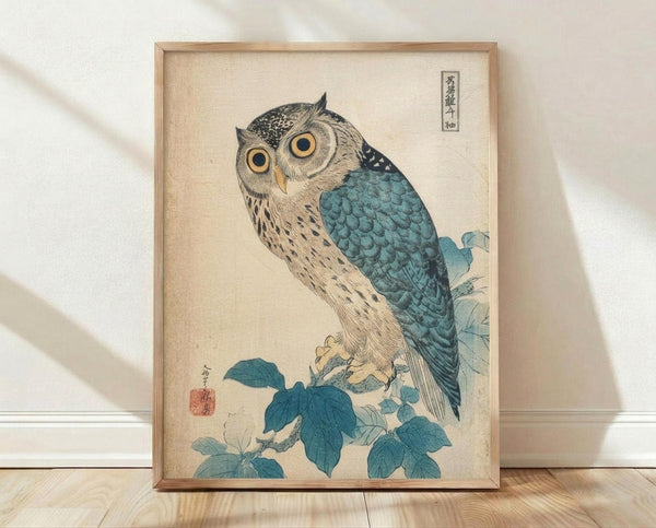 May include: Framed vintage Japanese woodblock print featuring an owl with large golden eyes perched on a branch with teal-blue leaves. The owl has detailed feather patterns in cream, brown, and blue tones against a beige background. Japanese text characters appear in the upper right corner with a red seal stamp on the left. The artwork is displayed in a light wooden frame, leaning against a white wall on light wood flooring with soft natural lighting.