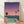 May include: Framed poster print leaning against white wall on wooden floor, showing retro-style illustration of Los Angeles cityscape at sunset with swimming pool in foreground, palm trees, airplane in purple-pink gradient sky, and sprawling city lights below in teal and coral color palette
