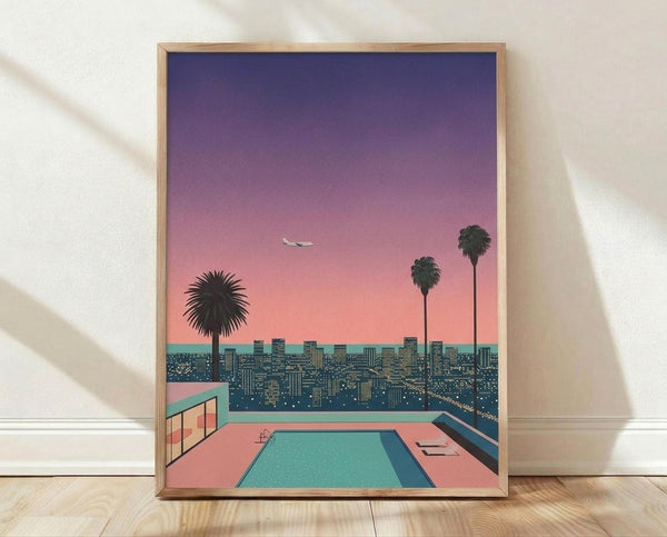 May include: Framed poster print leaning against white wall on wooden floor, showing retro-style illustration of Los Angeles cityscape at sunset with swimming pool in foreground, palm trees, airplane in purple-pink gradient sky, and sprawling city lights below in teal and coral color palette