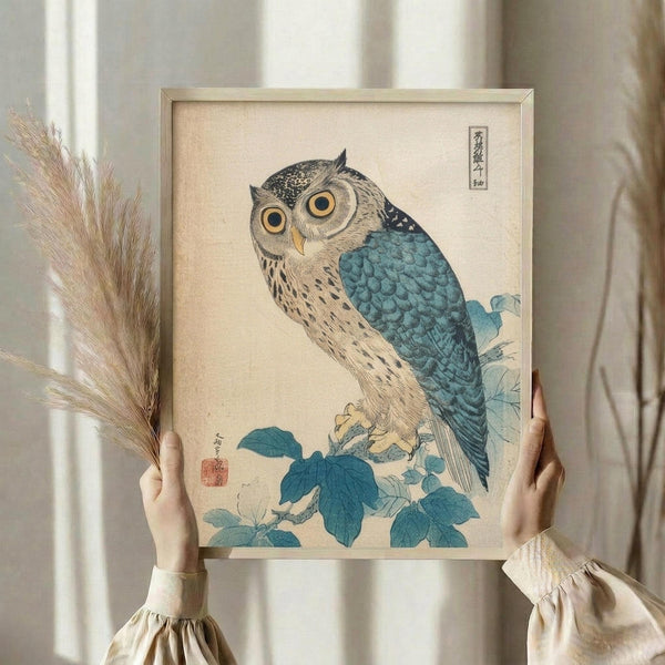 May include: Person holding framed Japanese woodblock print featuring owl with large golden eyes perched on blue-green foliage branch. Owl has cream and brown speckled feathers with teal-blue wings and tail. Traditional beige paper background with Japanese calligraphy in upper corner and red artist seal. Light wooden frame held against neutral curtained window with dried wheat stems visible on left side.