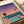 May include: Vintage-style poster print displayed on wooden surface beside white couch showing retro 1980s cityscape scene with swimming pool in foreground, palm trees, city skyline with illuminated buildings, gradient sunset sky transitioning from purple to pink, and small airplane silhouette, rendered in nostalgic Japanese-inspired aesthetic with turquoise pool water and coral pink tones