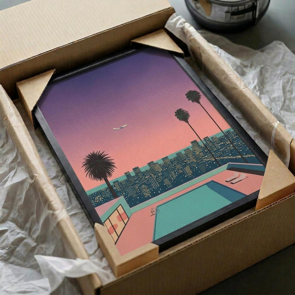 Framed art print being unboxed from cardboard packaging with white tissue paper. The artwork shows a retro-style cityscape at sunset with purple-pink gradient sky, palm trees, geometric swimming pool area in coral and teal colors, and urban buildings with lit windows. The print has a vintage 1980s aesthetic with bold colors and geometric shapes, displayed in a black frame surrounded by protective packaging materials.