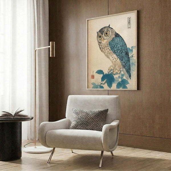 May include: Japanese woodblock print style artwork of an owl with blue and teal wings and cream colored body perched on flowering branch, displayed in light wooden frame on wood paneled wall above beige upholstered armchair with geometric patterned pillow, beside brass floor lamp and dark round side table with open book
