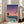May include: Framed poster print leaning against white wall next to beige sofa in bright room with large windows. Artwork shows retro-style illustration with gradient purple-to-pink sunset sky, three palm tree silhouettes, city skyline with lit buildings, and turquoise swimming pool in foreground. Print has light wood frame and vibrant 1980s aesthetic with coral, teal, and violet colors.