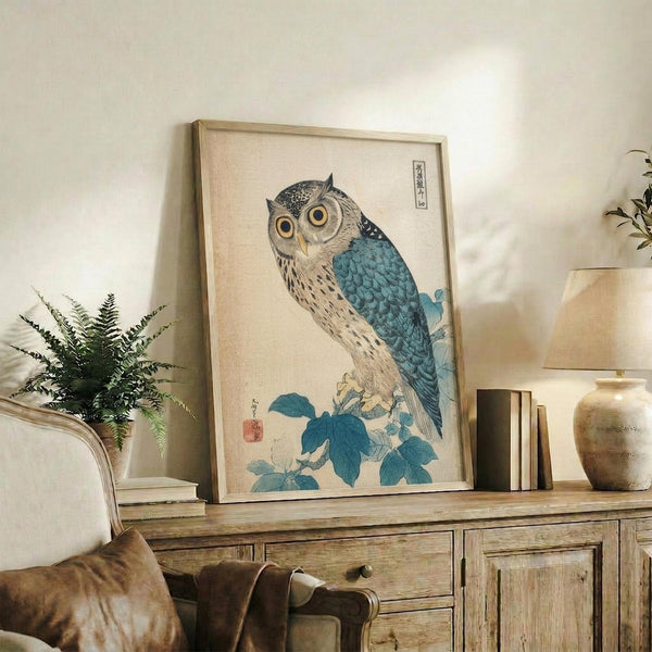 May include: Framed vintage Japanese woodblock print of an owl with blue wings and detailed feather patterns perched on flowering branch, displayed on rustic wooden sideboard alongside books, ceramic vases, and potted fern in neutral-toned living room setting.