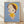 May include: Framed art print leaning against white textured wall on light wood floor. The artwork shows a stylized child-like character with an oversized head, brown bob haircut, yellow beret, and large green eyes against a soft blue background. The character wears a yellow dress with green button and has pale skin with rosy cheeks. Natural wood frame surrounds the print, with soft lighting creating gentle shadows on the wall.