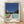 May include: Framed poster print leaning against white textured wall on light wooden floor with natural sunlight casting diagonal shadows. The artwork shows a modernist architectural scene with a swimming pool, contemporary house, palm tree, and deep blue sky in a clean geometric illustration style. Light wood frame surrounds the print which features the text "HIROSHI NAGAI" and "TOKYO 1973" along with colored dots in blue, peach, green, coral and navy.