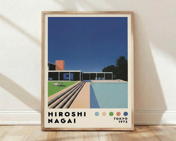 May include: Framed poster print leaning against white textured wall on light wooden floor with natural sunlight casting diagonal shadows. The artwork shows a modernist architectural scene with a swimming pool, contemporary house, palm tree, and deep blue sky in a clean geometric illustration style. Light wood frame surrounds the print which features the text "HIROSHI NAGAI" and "TOKYO 1973" along with colored dots in blue, peach, green, coral and navy.