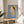 May include: Framed art print displayed on round wooden side table next to beige upholstered chair and plant. Artwork shows cartoon-style character with brown bob haircut, yellow beret and dress, holding small knife, set against blue background. Natural lighting creates soft shadows on white wall.