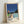 May include: Framed poster print leaning against white wall showing modern architectural scene with swimming pool, mid-century house, palm trees under deep blue sky. Features clean geometric lines in turquoise, coral, and green with "HIROSHI NAGAI" and "TOKYO 1973" text at bottom. Light wood frame, approximately 16x20 size, displayed on light wooden surface with natural lighting.