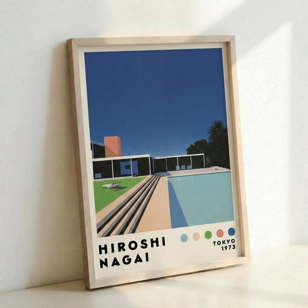 May include: Framed poster print leaning against white wall showing modern architectural scene with swimming pool, mid-century house, palm trees under deep blue sky. Features clean geometric lines in turquoise, coral, and green with "HIROSHI NAGAI" and "TOKYO 1973" text at bottom. Light wood frame, approximately 16x20 size, displayed on light wooden surface with natural lighting.