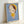 May include: Framed art print leaning against white wall showing cartoon-style character with oversized head, green eyes, brown hair with yellow headband, wearing yellow dress. Child-like figure rendered in soft colors against blue background. Light wood frame, appears medium-sized, photographed at angle showing depth and shadow on wall.