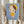 May include: Person in beige clothing holding framed art print against white curtained window, with dried wheat stems in left hand. The artwork shows a stylized child character with green eyes, brown and yellow hair, wearing yellow dress, holding small knife, set against blue background in light wooden frame.