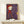 May include: Framed art print displaying a stylized character in red outfit and hat on purple watercolor background, shown in light wood frame on white shelf next to small wrapped gift with evergreen sprig