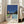 May include: Framed art print leaning against beige sectional sofa in bright white room with large windows. Print shows modernist poolside scene with swimming pool, contemporary house, palm trees against deep blue sky. Light wood frame, displayed on light hardwood floors next to sofa legs.