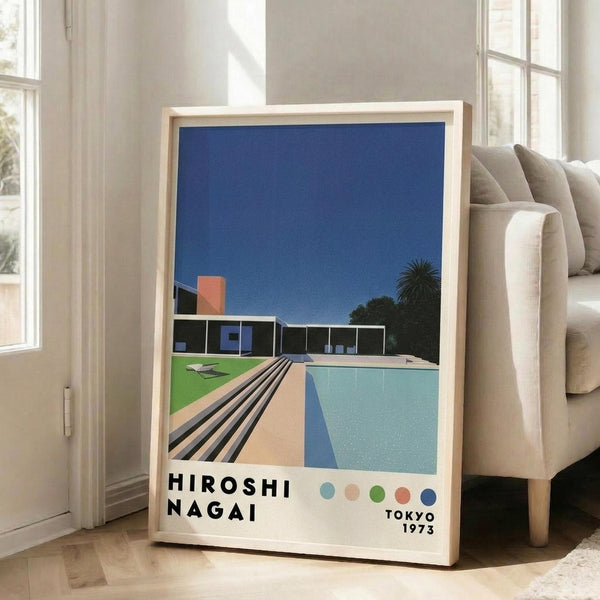 May include: Framed art print leaning against beige sectional sofa in bright white room with large windows. Print shows modernist poolside scene with swimming pool, contemporary house, palm trees against deep blue sky. Light wood frame, displayed on light hardwood floors next to sofa legs.