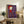 May include: Framed artwork displaying a stylized character in red hooded clothing on wooden stilts against a purple background, positioned on a rustic wooden sideboard alongside potted plants, books, and ceramic vessels in a cozy living room setting with neutral tones and natural lighting.