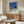 May include: Mid-century modern living room with wooden sideboard, white curved chair, and glass coffee table. Framed poster on wall shows modernist architecture with swimming pool, lawn, and blue sky in retro illustration style. Warm wood tones throughout room with natural lighting and minimalist decor including brass floor lamp and table lamp.