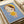 May include: Framed art print displayed on white bedding next to a beige pillow. The artwork features a stylized character with large green eyes, brown hair, and yellow halo against a blue background. The figure wears a yellow dress and holds a small white object. The print has a light wooden frame and shows a cute, anime-inspired illustration style with soft colors and simple shapes.