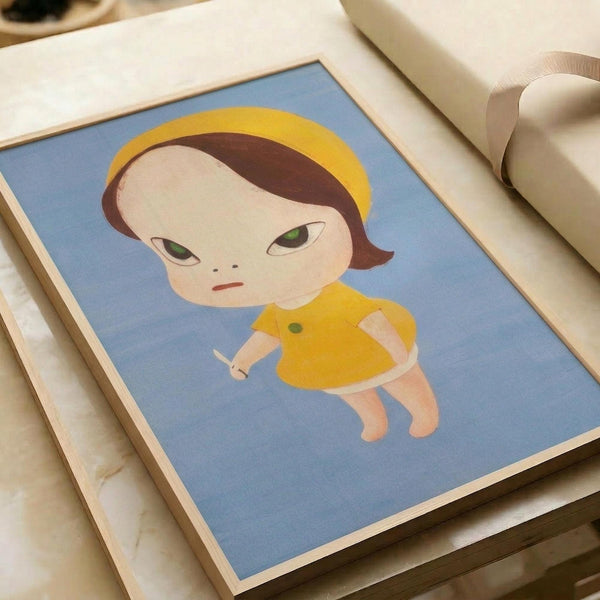 May include: Framed art print displayed on white bedding next to a beige pillow. The artwork features a stylized character with large green eyes, brown hair, and yellow halo against a blue background. The figure wears a yellow dress and holds a small white object. The print has a light wooden frame and shows a cute, anime-inspired illustration style with soft colors and simple shapes.
