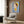 May include: Contemporary artwork featuring a stylized child character with large eyes, brown hair, and yellow dress against blue background, displayed in light wood frame on warm wood paneled wall. Modern beige armchair with geometric patterned pillow sits below, accompanied by brass floor lamp and dark side table with open book. Neutral curtains frame window on left.