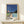 May include: Framed poster print featuring modernist architecture with swimming pool, geometric building, palm tree against deep blue sky. Art shows clean lines, retro color palette of blues, greens, coral pink. Print displayed on light wood shelf with small wrapped gift, natural lighting creates soft shadows on cream wall.