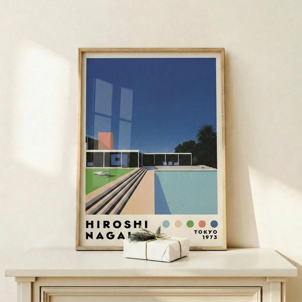 May include: Framed poster print featuring modernist architecture with swimming pool, geometric building, palm tree against deep blue sky. Art shows clean lines, retro color palette of blues, greens, coral pink. Print displayed on light wood shelf with small wrapped gift, natural lighting creates soft shadows on cream wall.