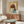May include: Modern living room with warm wood sideboard displaying framed artwork featuring cartoon-style girl with brown bob haircut and large yellow-green eyes on bright yellow background with collaged paper elements. White curved chair and glass coffee table visible in foreground, copper floor lamp stands nearby.
