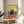 May include: Framed poster print featuring a black cat sitting on a coral-colored bar counter with wine glasses and bottle nearby, set against yellow and teal background. Print displayed on round wooden side table next to beige upholstered chair with decorative pillow, plant visible in corner, natural lighting creating window shadows on white wall.