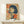May include: Framed vintage-style poster featuring a woman with black bob haircut and cigarette, rendered in beige, red, and blue colors with Japanese text, displayed on light wood console table beside small white wrapped gift with dried grass accent against white wall